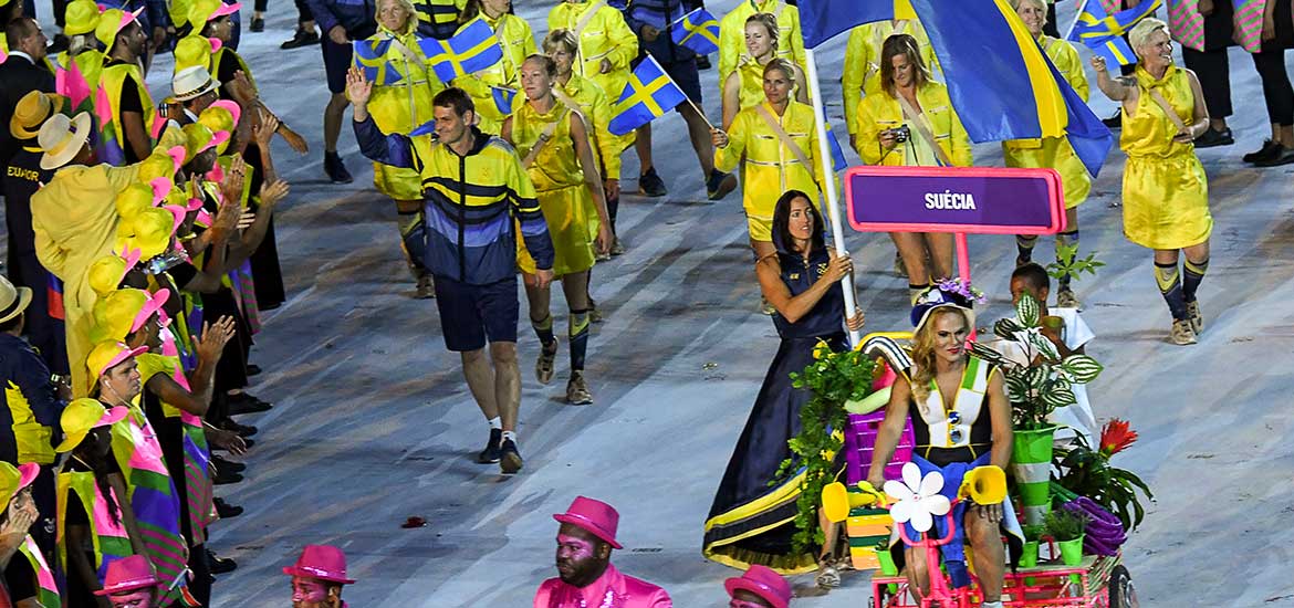 En trupp av människor i gula och blå kläder. Längst fram en kvinna i blå klänning med gula detaljer som bär på en svensk fana.