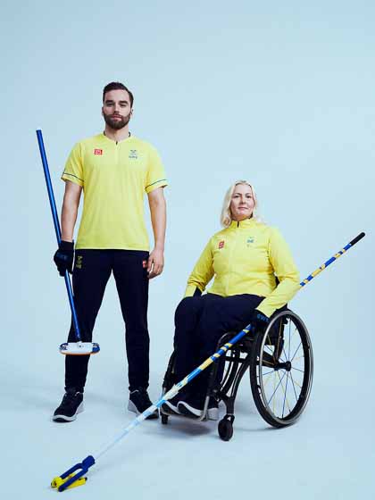 Två curlingspelare, en man som står och en kvinna i rullstol. Båda har citrongula kläder på överkroppen och mörkblå byxor.