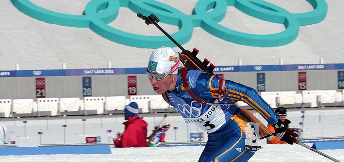 En skidskytt i full fart med skidskyttevallen i bakgrunden och de olympiska ringarna ovanför. 