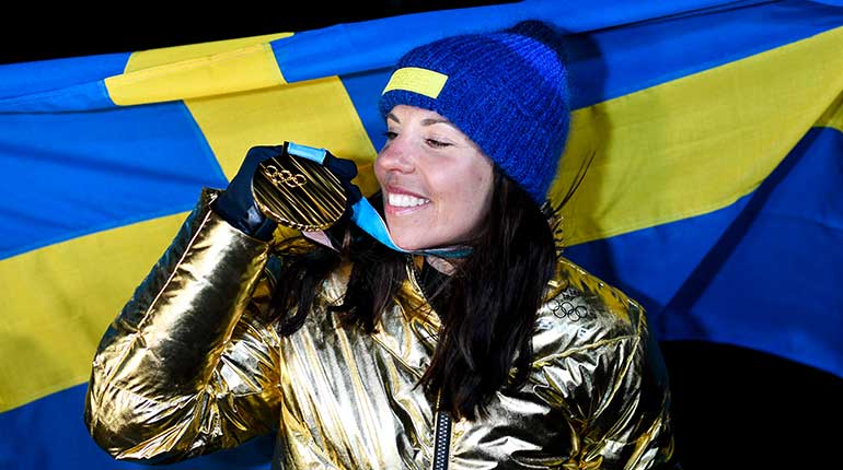 Charlotte tittar på sin guldmedalj med en dunjacka av guld och svenska flaggan i bakgrunden. 