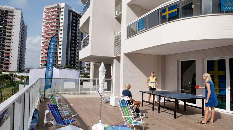 En terass med solstolar och vita hus i bakgrunden. Tre tjejer spelar pingis på terrassen iklädda svenska truppkläder. 
