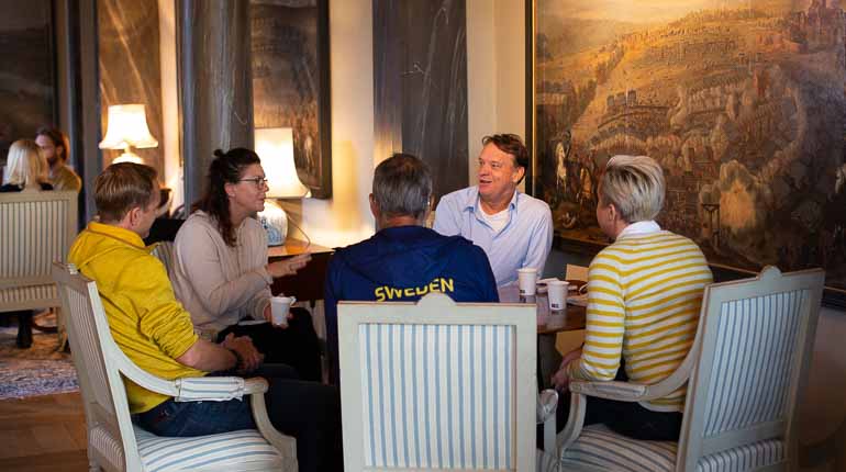 En grupp personer sitter runt ett bord och pratar med antika möbler och tavlor.