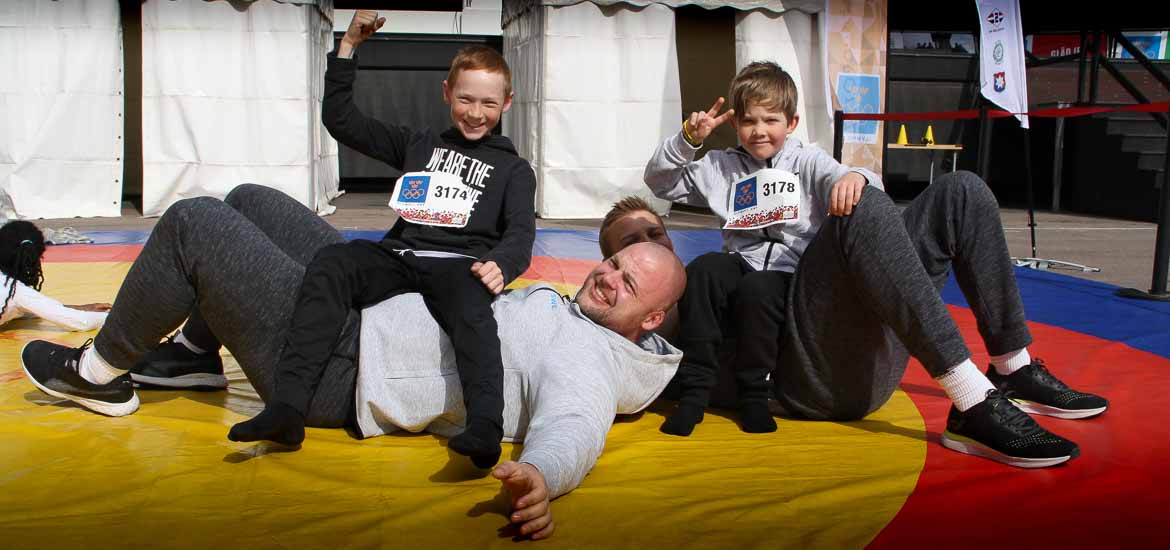 Två leende barn sitter ovan två storvuxna brottare och gör segertecken. 